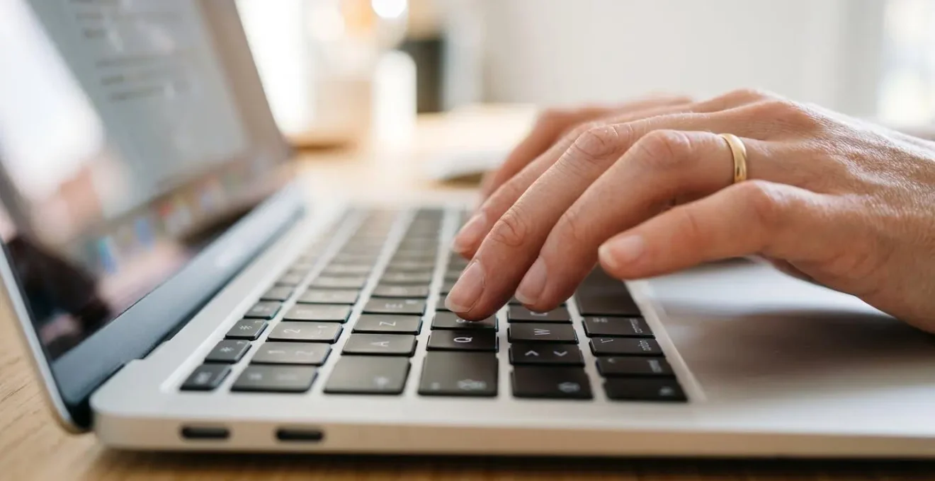 Main féminine tapant sur le clavier d'un MacBook moderne avec écran totalement flouté en arrière-plan sur un bureau épuré blanc et bois clair
